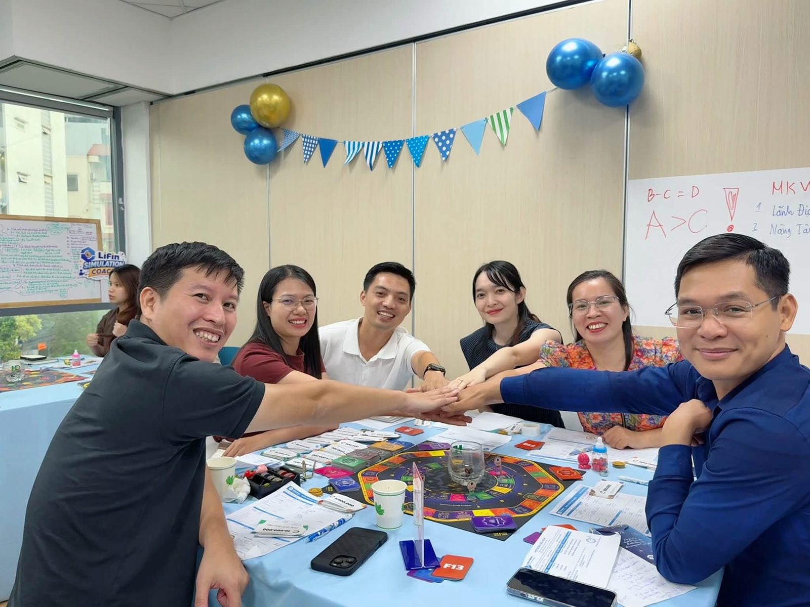 Nhóm người chơi cùng đặt tay chồng lên nhau quanh bàn LiFin Game, cười rạng rỡ sau buổi chơi